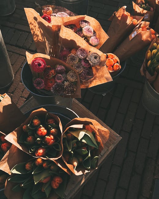A selection of flower bouquets and arrangements placed outdoors on wooden tables and metal containers, wrapped in brown kraft paper. The bouquets feature an array of vibrant colors, predominantly reds