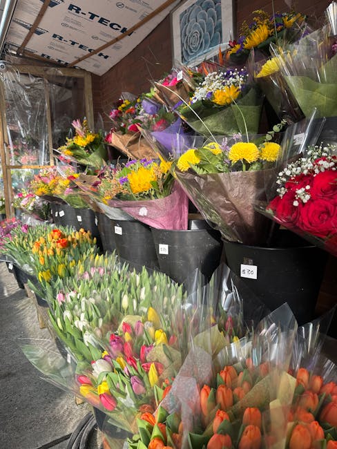 An indoor display of vibrant flower bouquets arranged in black buckets and plastic wrapping, featuring a variety of fresh blooms including yellow chrysanthemums, red roses, pink and white tulips, oran