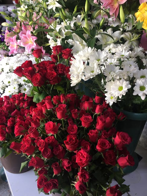 An arrangement of fresh flowers including a large bouquet of red roses with lush green stems, surrounded by smaller clusters of white daisies, pink lilies, and yellow blooms. The flowers are displayed