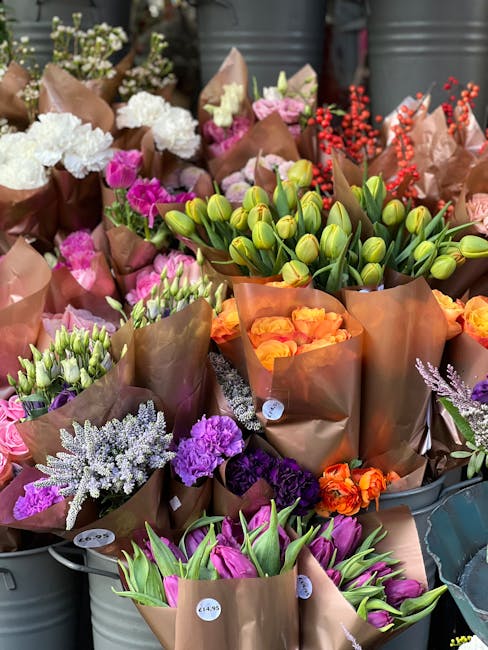 A vibrant display of freshly cut flowers arranged in brown paper wrapping, including white and purplestatice, pink and purple tulips, orange and pink roses, and green tulip buds, showcasing a variety