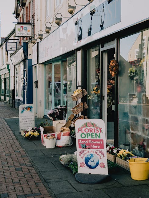 The exterior of a flower shop with large glass windows displaying vibrant floral arrangements inside. Outside, a variety of potted chrysanthemums and scattered bouquets of mixed flowers are arranged o