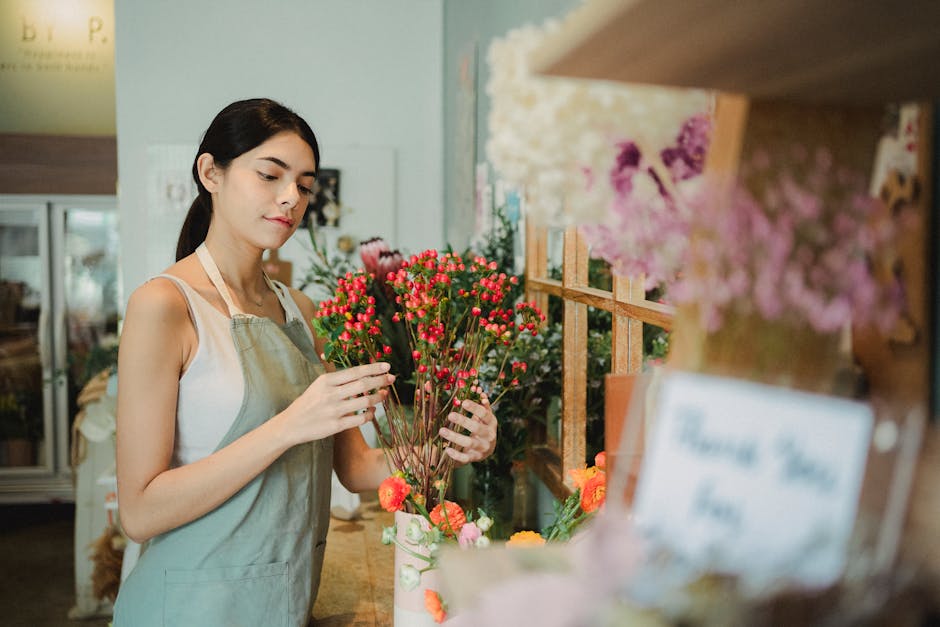A young woman with dark hair wearing a light-colored apron is selecting or arranging a bouquet of small, bright pink and red flowers with green stems inside a florist shop. She stands next to a wooden