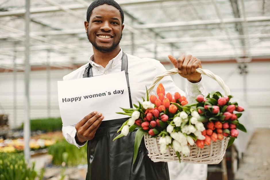 A cheerful male florist inside a greenhouse or floral shop, holding a white wicker basket filled with fresh tulips in shades of pink, red, and white, interspersed with green leaves. He is smiling and 